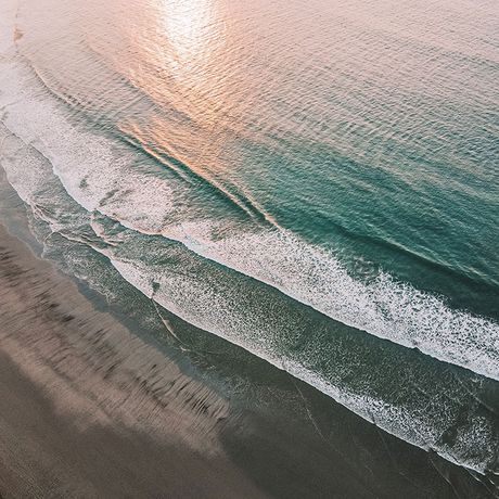 Das Meer bricht mit feinen Schaumkronen an einen naturbelassenen Strand während Sonnenlicht leicht auf dem Wasser reflektiert.