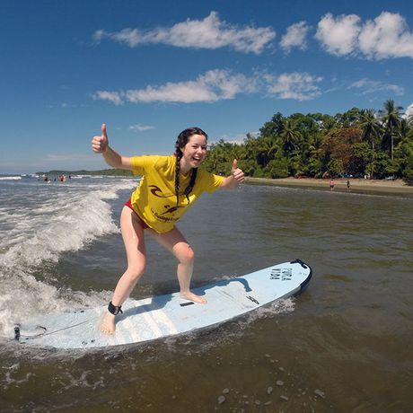 EIne junge Frau steht mit Daumen hoch auf dem Surfbrett und lässt sich von einer Welle in Richtung Strand tragen. 