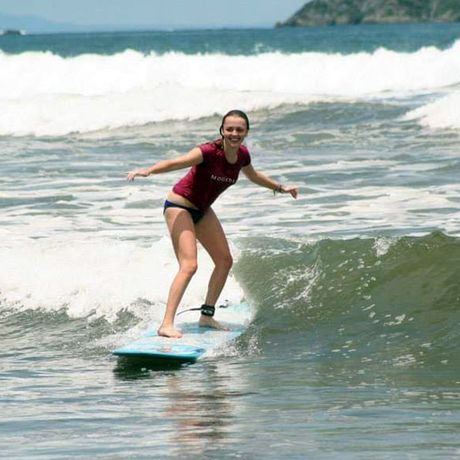 Eine junge Frau surft auf einer Welle und lächelt vor lauter Freude und Spaß über das ganze Gesicht.